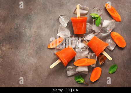 Succo di carota congelato su bastone, sfondo marrone scuro. Sano dessert vegano estivo. Gelati. Vista dall'alto, disposizione piatta, spazio di copia Foto Stock