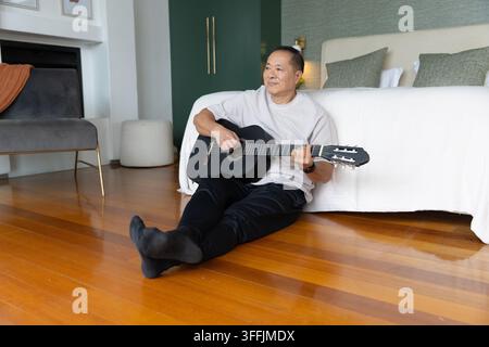 Uomo cinese anziano seduto sul pavimento di legno in camera da letto con una chitarra acustica nera, appoggiato all'indietro Foto Stock
