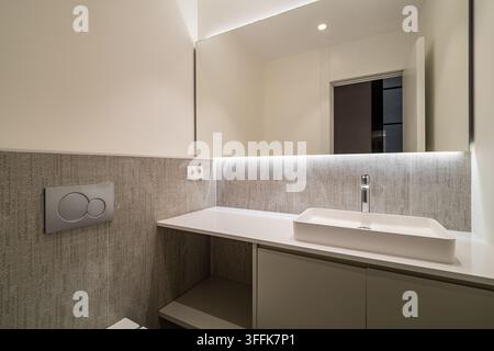 Interno di bagno contemporaneo con lavandino rettangolare bianco, mobiletto vanità minimalista, piastrelle murali texture, illuminazione calda e soffusa, finiture neutre Foto Stock