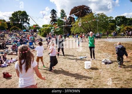 SCENA VIBRANTE DI FESTIVAL CON FAMIGLIE E BOLLE GIGANTI, REPORTAGE, GREEN MAN FESTIVAL 2014, BRECON, WALES, UK, 2014: Una fotografia vivace e grandangolare cattura la gioia di un festival estivo, con bambini che inseguono bolle colorate mentre la folla si rilassa su una collina erbosa. Foto: Rob Watkins. INFO: Il Green Man Festival si tiene ogni anno ad agosto nelle Brecon Beacons. Celebra musica alternativa, arte, letteratura, scienza e una cultura a misura di famiglia in uno splendido paesaggio montano. L’evento del 2014 è stato caldo e soleggiato. Foto Stock