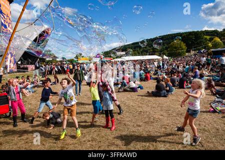 BAMBINI ECCITATI CHE INSEGUONO UNA CASCATA DI BOLLE, REPORTAGE, FESTIVAL GREEN MAN 2014, BRECON, WALES, UK, 2014: Un gruppo di bambini estatici salta e cerca di raggiungere una cascata massiccia di bolle iridescenti che fluttuano nell'aria durante un festival all'aperto soleggiato. Foto: Rob Watkins. INFO: Il festival Green Man si tiene ogni anno ad agosto nelle Brecon Beacons. Celebra musica alternativa, arte, letteratura, scienza e cultura a misura di famiglia in uno splendido scenario montano. L'evento del 2014 è stato caldo e soleggiato. Foto Stock