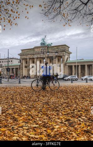 Vista del Portone di Brandenburgo in autunno con un ciclista in giacca blu in piedi su un sentiero coperto di foglie cadute a Berlino, Berlino, Germania. Foto Stock