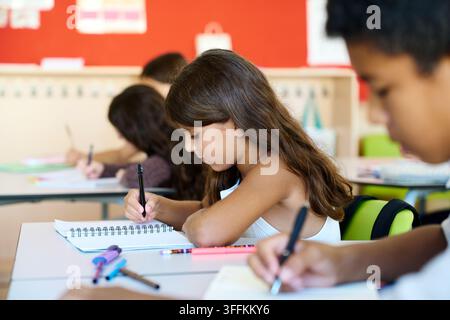 Bambini seduti a banchi in una classe luminosa, concentrati sulla scrittura e sull'apprendimento, rappresentando il ritorno all'istruzione scolastica Foto Stock