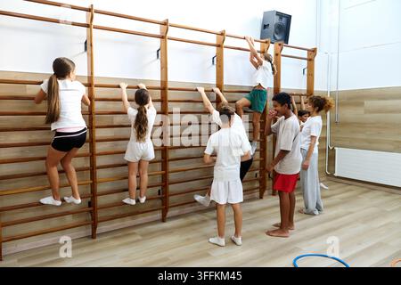 Diversi studenti impegnati nell'educazione fisica, arrampicandosi e aggrappandosi alle sbarre murali in una palestra scolastica. Praticano sport e fitness Foto Stock