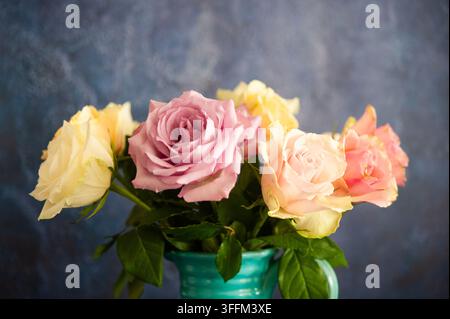 Mazzo di rose pastello rosa, crema e gialle in un vaso turchese su uno sfondo blu-grigio morbido, fotografato come natura morta floreale Foto Stock