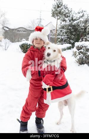 Ragazzo in un pigiama da neve rosso abbraccia il suo cane vestito con una giacca di Babbo Natale mentre cammina in un parco coperto di neve Foto Stock