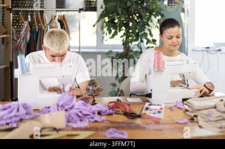 I sarti stanno lavorando su una collezione di abiti. Sarto maschio che lavora in squadra con sarte asiatiche, cuciono il campione per l'approvazione con cust Foto Stock