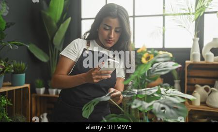 Donna fiorista in grembiule tiene uno smartphone con una mano e tocca una foglia di pianta con l'altra mano in uno studio; messa a fuoco calma. Foto Stock