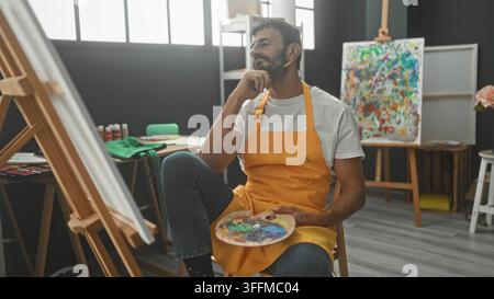 Pittore uomo con grembiule giallo tiene una tavolozza e appoggia la mano sul mento mentre è seduto all cavalletto nello studio; contemplazione creatività. Foto Stock