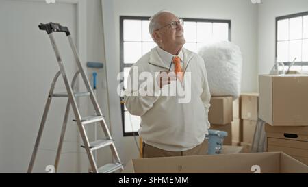 Uomo anziano che tiene la penna al mento tra le scatole di cartone e la scala all'interno di un edificio, indossando occhiali e cravatta arancione; pianificazione nostalgica. Foto Stock