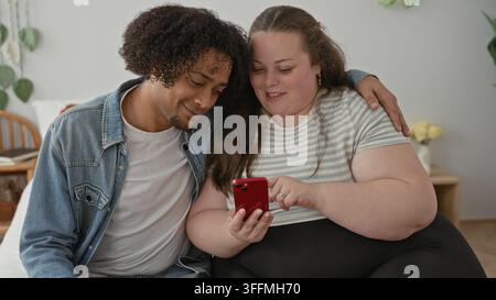 Uomo e donna più grande siedono sul letto, la donna tiene lo smartphone rosso e l'uomo mette il braccio intorno alla spalla all'interno dell'edificio; intimità e gioia. Foto Stock