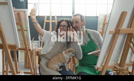 Una donna di mezza età solleva lo smartphone per un selfie in uno studio d'arte con un uomo che dipinge su un cavalletto e un piccolo cane in grembo; gioia di compagnia. Foto Stock