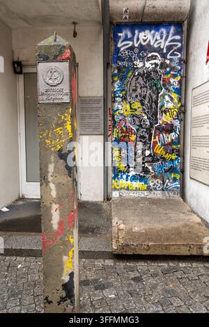 Segmento del muro di Berlino coperto di graffiti accanto a un posto di confine della DDR con emblema di stato su ciottoli, Berlino, Germania Foto Stock