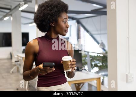 Donna adulta africana in piedi in ufficio con una maglia marrone, tiene un telefono e un bicchiere da asporto e guarda a destra Foto Stock