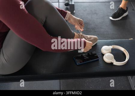 Compagne di palestra femminili sedute su una panchina, una che allaccia i lacci delle scarpe beige in una maglia marrone accanto al telefono Foto Stock