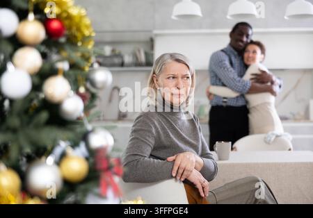 Una vecchia triste donna seduta al tavolo della cucina con le spalle alla coppia che posa felicemente in cucina con l'albero di Natale Foto Stock