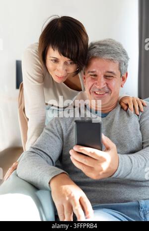 Coppia matura sorridente si rilassa a casa mentre l'uomo tiene uno smartphone e la donna si appoggia vicina, condividendo un momento caloroso e affettuoso mentre guardano lo scr Foto Stock