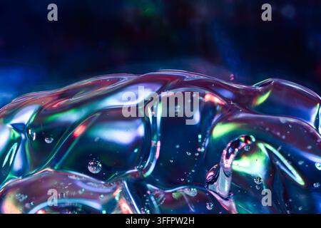 Vista macro di una superficie di gel liquido iridescente e lucida, con pieghe traslucide, piccole bolle e riflessi vividi in blu, rosa, verde e viola a Foto Stock