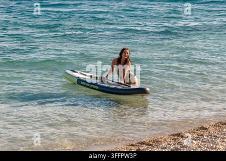 Una donna sorridente si bilancia su una pagaia in acque basse vicino a una riva di ciottoli. Foto Stock