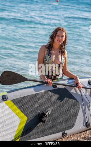 Una donna sorridente in costume da bagno a fantasia si trova con una pagaia su una tavola da paddle vicino alla riva. Foto Stock
