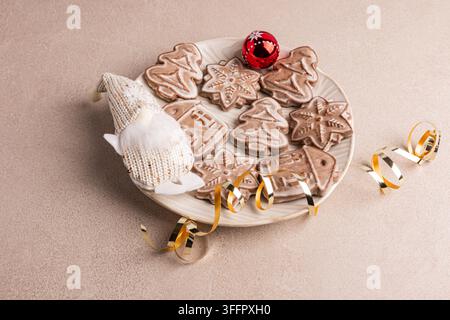 Biscotti di zenzero natalizi fatti in casa in forme festive disposti su un piatto di ceramica con un gnomo lavorato a maglia, un ornamento rosso e un nastro dorato su uno sfondo rustico Foto Stock