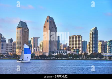 Un'illuminante barca a vela scivola attraverso la baia davanti alla suggestiva skyline di San Diego. Il lussureggiante Coronado Park incornicia il lungomare sotto una limpida California Foto Stock