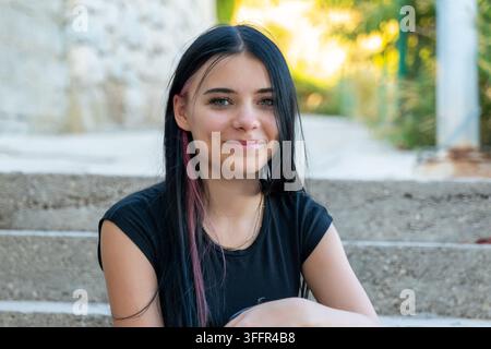 Una giovane donna sorridente con lunghi capelli neri e una ciocca rosa è seduta all'aperto. Foto Stock