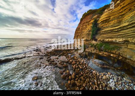 Scogliere di arenaria stratificate si ergono sopra una spiaggia rocciosa alle Sunset Cliffs a San Diego in California mentre le onde dell'Oceano Pacifico si infrangono sotto un cielo drammatico Foto Stock