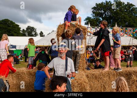 DIVERTIMENTO FAMILIARE SUI BALLI DI FAGIOLO A UN FESTIVAL ESTIVO, REPORTAGE, FESTIVAL GREEN MAN 2015, BRECON, GALLES, REGNO UNITO, 2015: Un uomo con cappello e maglietta si trova in mezzo a una scena caotica e gioiosa di numerosi bambini che si arrampicano e giocano su una grande pila di balli di fieno in un campo soleggiato. Foto: Rob Watkins. INFO: Il festival Green Man si tiene ogni anno nelle Brecon Beacons ad agosto. Celebra musica alternativa, arti, letteratura, scienza e cultura a misura di famiglia in uno splendido scenario montano. L'evento del 2015 è iniziato soleggiato ma è diventato fangoso a causa della pioggia caduta durante la notte negli ultimi due giorni. Foto Stock