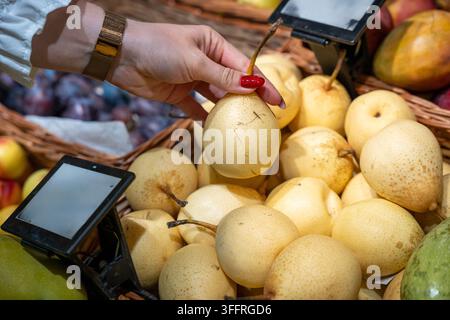 Donna adulta solleva con cura una pera dal cestino del mercato mentre i pannelli digitali dei prezzi incorniciano prodotti freschi. Gesto della cliente e manicure rossa Foto Stock
