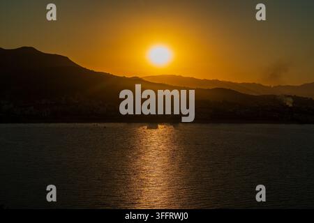 Sole dorato brillante che tramonta sulle vette delle montagne costiere con una scia di luce scintillante riflessa sull'acqua calma del mare, costa mediterranea, Marocco. Foto Stock