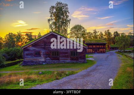 Casa tradizionale in tronchi ed edifici rurali al tramonto nel museo all'aperto di Maihaugen, Lillehammer, Norvegia, con cielo dorato e alberi circostanti. Foto Stock