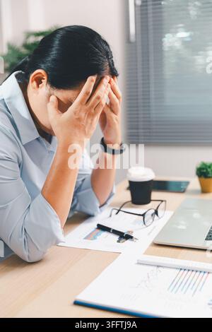 Donna d'affari stressata che copre il viso mentre lavora su un laptop in un ufficio moderno, mostrando burnout, ansia, affaticamento, pressione lavorativa, scadenza Foto Stock