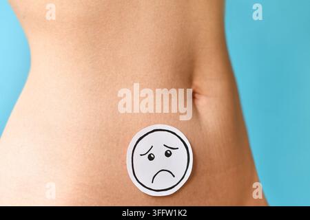 Faccia triste sull'addome di una donna che simboleggia dolori mestruali, endometriosi, problemi di fertilità e problemi di salute riproduttiva davanti a uno sfondo blu Foto Stock