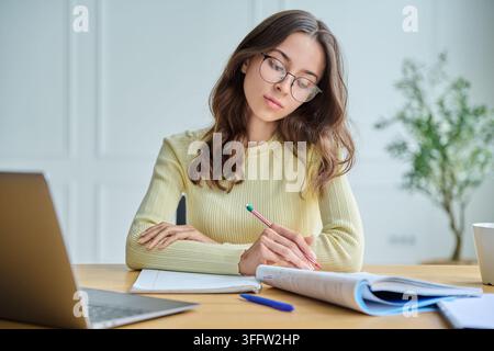 Studentessa concentrata che prende appunti alla scrivania con vista frontale sul laptop Foto Stock