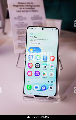 RIGA, LETTONIA. 25 agosto 2026. Primo piano dello smartphone Samsung Galaxy S26 FE visualizzato su un supporto, che mostra l'interfaccia della schermata principale con il lettone Foto Stock