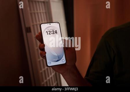 RIGA, LETTONIA. 25 agosto 2026. Persona che tiene lo smartphone Samsung Galaxy S26 FE in un ambiente scarsamente illuminato, mostrando lo schermo di blocco con l'ora Foto Stock