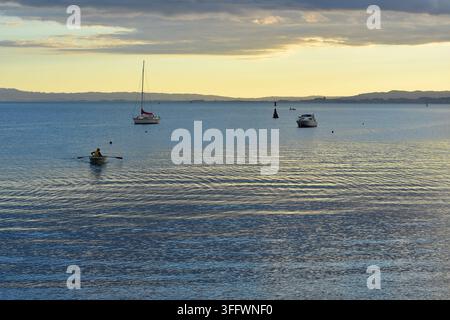 Scena costiera tranquilla con una persona che rema in un piccolo dinghy su acque blu calme, con barche ormeggiate e un dolce tramonto all'orizzonte. Foto Stock