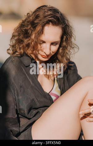 Una giovane donna con capelli castani ricci che posa all'aperto in una maglietta nera e un costume da bagno fantasia, godendosi la calda luce del sole in vacanza al mare Foto Stock