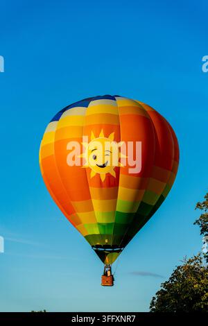 Bakewell, Regno Unito - 05 settembre 2026: Vista di un pallone aerostatico colorato con un design a faccia di sole che fa l'occhiolino, che galleggia contro un cielo blu terso. Foto Stock