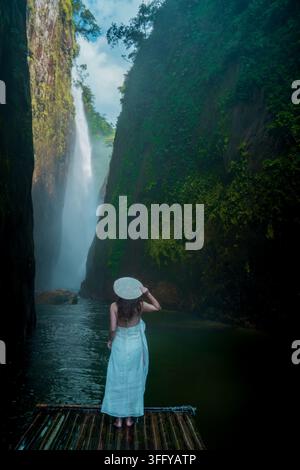 Donna in vestito bianco in piedi su una zattera di bambù di fronte a una cascata alta, Muong Hum, Lao Cai, Vietnam Foto Stock