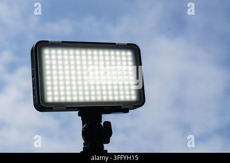 Luce a pannello LED brillante su un piccolo supporto contro un cielo blu nuvoloso e morbido, che mostra attrezzature per l'illuminazione esterna per video, fotografia e studio. Foto Stock