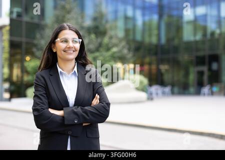 Giovane donna imprenditrice indiana in abito nero e occhiali, in piedi all'aperto con le braccia incrociate accanto a un edificio di uffici in vetro, sorride con sicurezza e incarna leadership e ambizione Foto Stock