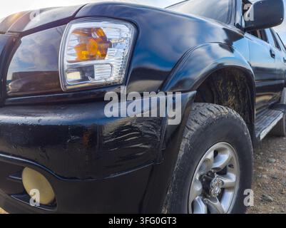 Vista bassa dettagliata dell'angolo anteriore di un SUV nero parcheggiato su una superficie di terra. Il pneumatico robusto e il paraurti consumato indicano un uso fuoristrada, con riflessi del cielo sulla carrozzeria lucida. Foto Stock