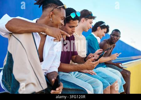 Diversi giovani amici maschi seduti all'aperto, connettendosi con dispositivi digitali e interagendo mentre utilizzano i loro telefoni mobili Foto Stock