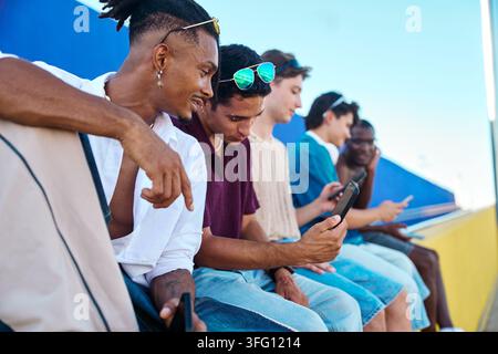 Gruppo diversificato di giovani che interagiscono con i loro smartphone mentre sono seduti insieme all'aperto, rappresentando la connessione digitale e lo stile di vita giovanile Foto Stock
