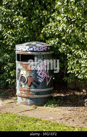Un bidone della spazzatura arrugginito e coperto di graffiti si trova accanto a una fitta siepe su un sentiero a King’s Gardens, Southport, Regno Unito. Foto Stock