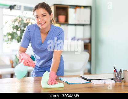 Ritratto di una donna delle pulizie che lavora in un ufficio con attrezzature per la pulizia e spolvera una scrivania Foto Stock