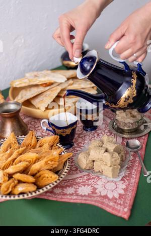 Spread di caffè e dolci mediorientali. Una mano versa il caffè da una caraffa vintage blu cobalto in una tazza. Dolci sul tavolo: baklava al miele e halva su una tovaglia rossa a motivi. Foto Stock