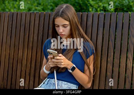 Giovane donna che scrive sul cellulare seduta all'aperto. Giovane donna utilizza uno smartphone seduta su una panchina di legno in un parco con fogliame verde nell' Foto Stock